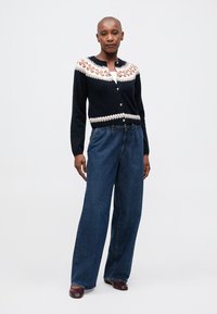 Cardigan marine avec motif floral dans des tons de marron et crème, présentant des boutons sur le devant. Associé à un jean bleu à jambes larges et des chaussures bordeaux.