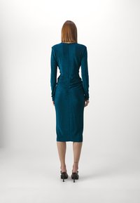 Turkos långärmad klänning med en figursydd silhuett. Har Rynkning bak och en knälång nederkant. Slät, glänsande tygtextur.