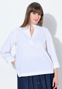 Camicia bianca a maniche lunghe con colletto senza bottoni e vestibilità leggermente ampia. Presenta un orlo dritto e una texture liscia, abbinata a una gonna scura.