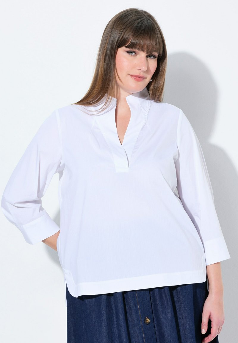 Camicia bianca a maniche lunghe con colletto senza bottoni e vestibilità leggermente ampia. Presenta un orlo dritto e una texture liscia, abbinata a una gonna scura.