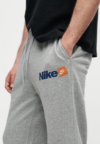 Άνδρας που φοράει γκρι φόρμες Nike με το ένα χέρι στην τσέπη, μαύρο πουκάμισο μερικώς ορατό, πάνω σε απλό φωτεινό φόντο.
