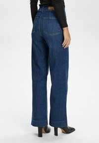 High-waisted, wide-leg dark blue denim jeans with back pockets, finished with contrasting stitching. Worn with black heeled shoes.