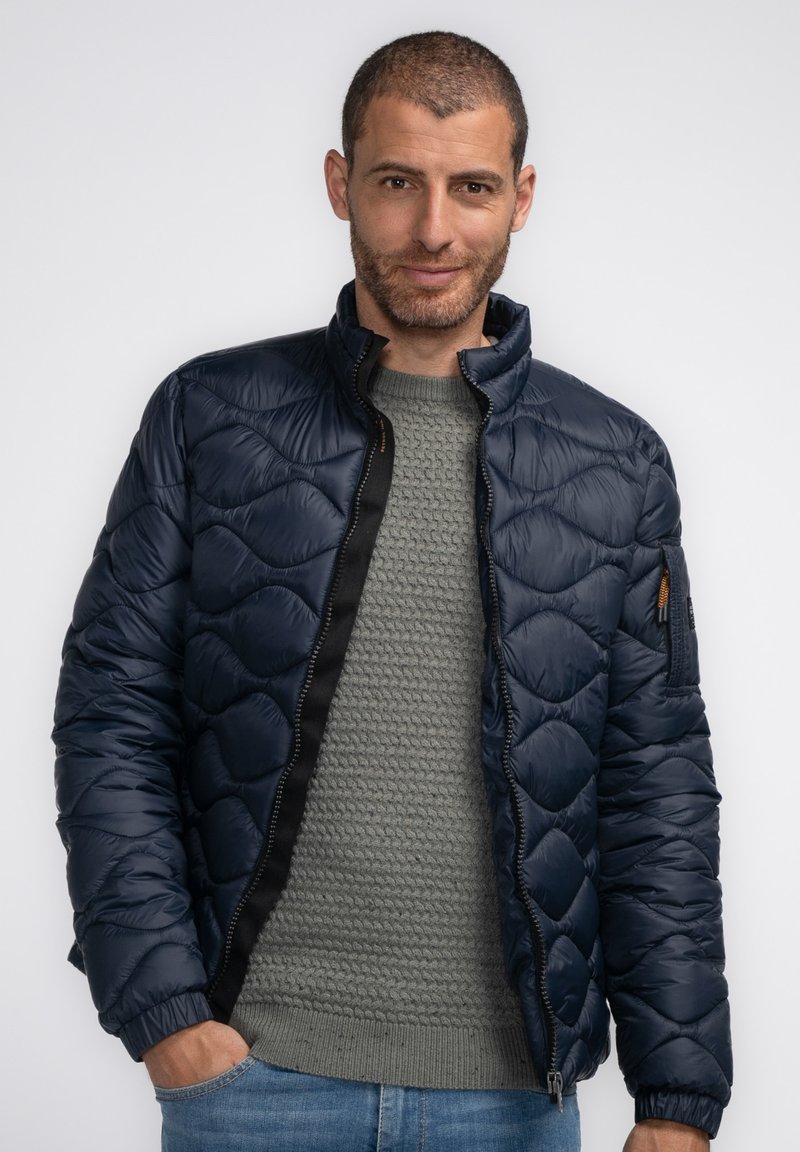 Marineblaues gestepptes Jacket mit Stehkragen, Reißverschluss und seitlichen Taschen, getragen über einem hellgrauen Strickpullover. Mit schwarzen Reißverschlussdetails.