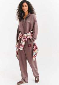 Brązowa bluza z kapturem i pasujące spodnie dresowe, z kołnierzem na suwak do ćwiartki. Koszula w kratę związana w talii, z czerwonymi, białymi i niebieskimi tonami.
