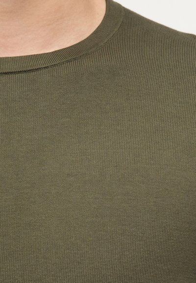 Κοντινό πλάνο ενός ατόμου που φοράει πουλόβερ ελιάς με ριμπ πλέξη και στρογγυλή λαιμόκοψη.