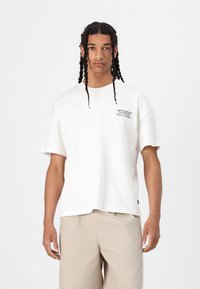Witte katoenen t-shirt met korte mouwen, een relaxte pasvorm en een bedrukte logo op de linkerborst. Soepele textuur, minimalistisch ontwerp.