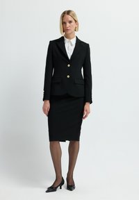 Schwarzer, maßgeschneiderter Anzug mit einer taillierten Blazerjacke, goldenen Knöpfen und passendem Bleistiftrock. Über einem weißen, kragenlosen Hemd getragen. Schwarze High Heels runden den Look ab.