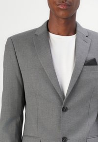 Grå skreddersydd blazer i strukturert stoff, med spiss krage, to knapper, og en brystlomme med svart aksent. Båret over en hvit skjorte.