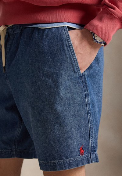 Κοντινό πλάνο προσώπου που φοράει μπλε τζιν σορτς με κόκκινο λογότυπο αλόγου, χέρι στην τσέπη, κόκκινο φούτερ και ρολόι με μπλε και λευκό λουράκι.