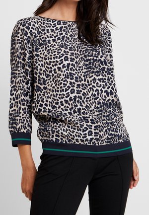 Kvinde iført en beige og sort leopardmønstret bluse med navy ærmekanter og kant med en grøn stribe, kombineret med sorte bukser.