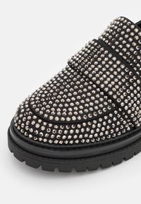 Chaussure mocassin noire avec semelle épaisse recouverte de petits clous argentés répartis uniformément sur la surface supérieure.