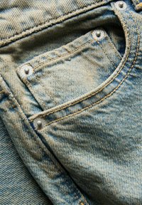 Denim fabric close-up featuring a light blue color, visible texture, and reinforced stitching with snap buttons on a pocket edge.