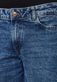 Mėlyni džinsai su klasikinio penkių kišenių dizainu, varinėmis sagomis ir kontrastingais siūlais, turintys nusidėvėjusį tekstūrą ir šiek tiek išblukusią spalvą.