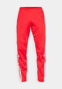 Červené sportovní kalhoty Adidas s bílými bočními pruhy, zapínacími kapsami, elastickým pasem a nášivkami s logem Adidas na nohavicích na bílém pozadí.