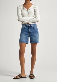 Suéter ligero con cuello redondo y cierre de botones, combinado con pantalones cortos de mezclilla azules y sandalias planas negras con tiras y acentos de tachuelas.