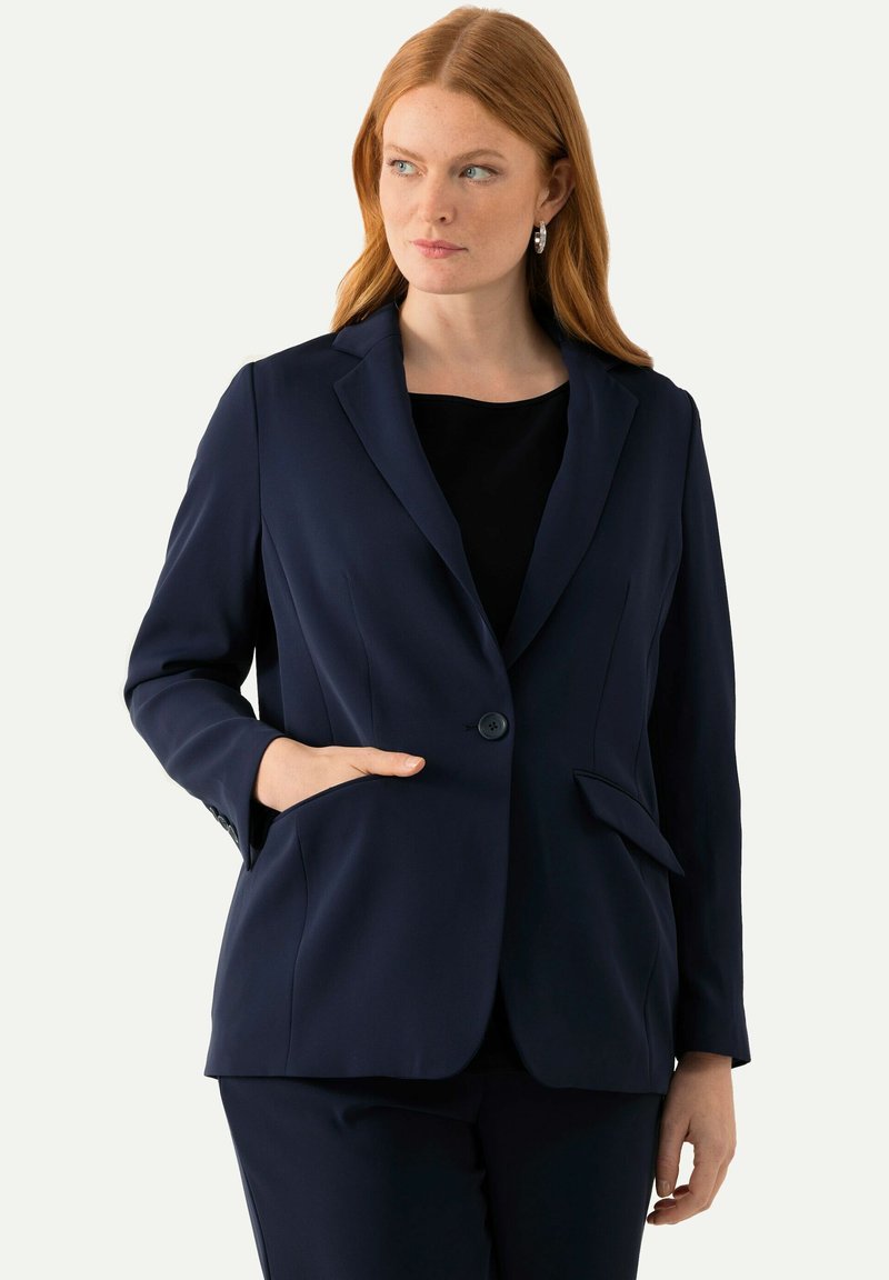 Blazer blu navy, design su misura, chiusura con un bottone, due tasche frontali, tessuto liscio, vestibilità aderente, indossato con una maglia nera.