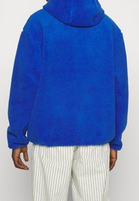 Pullover in pile blu con colletto alto e maniche lunghe, caratterizzato da una superficie texturizzata. Abbinato a pantaloni bianchi a righe.
