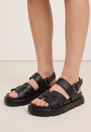 Pies con sandalias negras de suela gruesa con correas ajustables sobre los dedos y alrededor de los tobillos, de pie sobre un suelo claro.