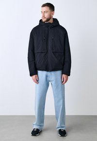 Chaqueta negra acolchada con capucha, que presenta dos bolsillos frontales y detalles en cremallera. Combinada con jeans de color azul claro y zapatillas negras.