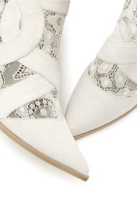 Bottes blanches à bout pointu et cheville avec panneaux en dentelle beige et superpositions en daim, vues de dessus sur un fond blanc.