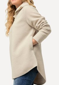 Giacca in fleece beige con cappuccio, dotata di un colletto alto, tasche laterali e un orlo posteriore più lungo. Leggera e con una texture morbida.