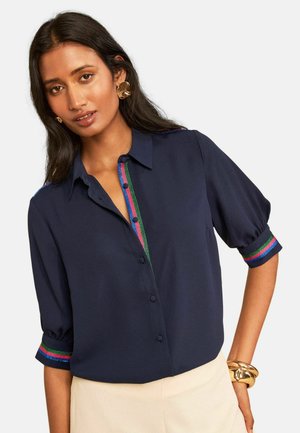Pikkade tumedate juustega naine kannab tumesinist pluusi, mille varrukatel ja nööpide ribal on värvikas triibuline ääris, sobitatud heledate pükstega.