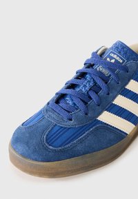 Adidaș bleu cu panouri din suede, dungi bej deschis, șireturi albastre și o talpă din cauciuc translucid, surprins pe un fundal alb.