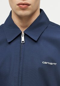 Κοντινό πλάνο ενός ατόμου που φοράει μπλε σκούρο ζακέτα με φερμουάρ, με μυτερό γιακά και λευκό λογότυπο "Carhartt" στο στήθος.