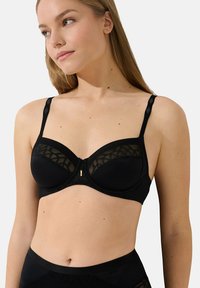 Soutien-gorge noir avec panneau en maille, support à armatures, bretelles réglables et un détail doré à l'avant. Texture de tissu lisse.