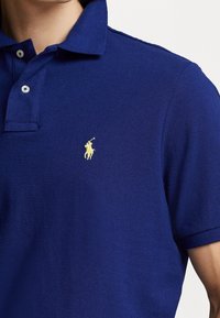 Námořnická polo košile vyrobená z texturované tkaniny; má žluté vyšívané logo na levém prsu a dvouknoflíkovou légu s bílými knoflíky.