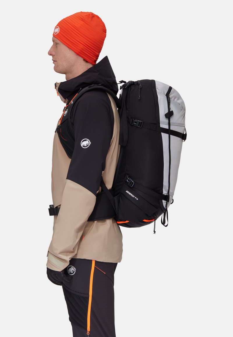 Svart och grå skidryggsäck med orange detaljer, vattentätt material, justerbara remmar och rullstängning. Bärs av en person i friluftskläder.