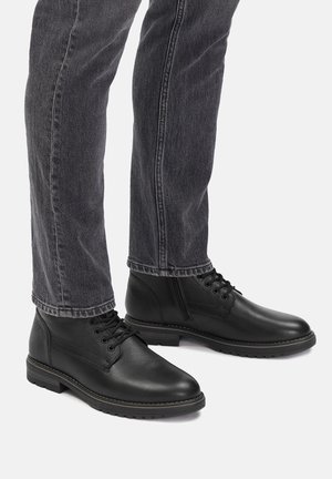 Czarne skórzane botki za kostkę z sznurówkami, o zaokrąglonym nosku, teksturowanej powierzchni i gumowej podeszwie. Noszone z szarymi dżinsami.