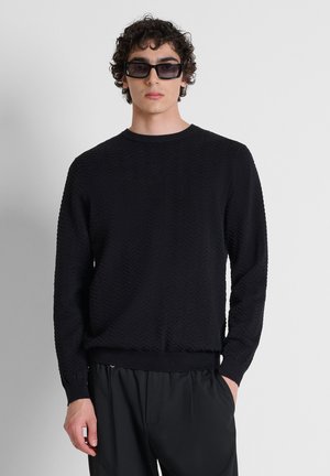 Junger Mann mit lockigem Haar, der große schwarze Sonnenbrillen, einen strukturierten schwarzen Pullover und schwarze Hosen trägt und vor einem schlichten hellen Hintergrund steht.
