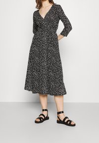 Czarna sukienka z dopasowanym gorsetem, rękawami o długości trzech kwart, oraz średniej długości rozkloszowaną spódnicą; ozdobiona białym, plamkowanym wzorem. Czarne sandały na ramiączkach.