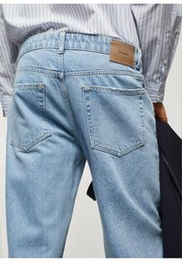 Los jeans de denim azul claro presentan un diseño de cinco bolsillos, un parche de cuero en la parte posterior y costuras estándar. La tela parece suave y ligeramente texturizada.
