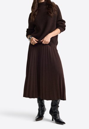 Femme portant un pull côtelé marron foncé, une jupe midi plissée assortie, des bottes en cuir noires à talons hauts, et des bijoux en argent au poignet et au doigt.