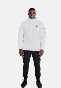 Giacca imbottita bianca con un emblema rosso sul petto, pantaloni cargo neri e sneakers nere, in piedi di fronte a uno sfondo semplice.