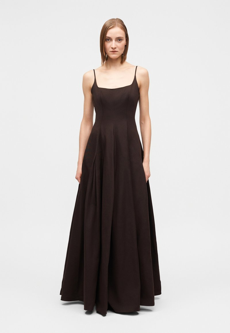 Mulher vestindo um vestido preto longo até ao chão, sem mangas e com alças finas, em pé contra um fundo cinza claro e liso.