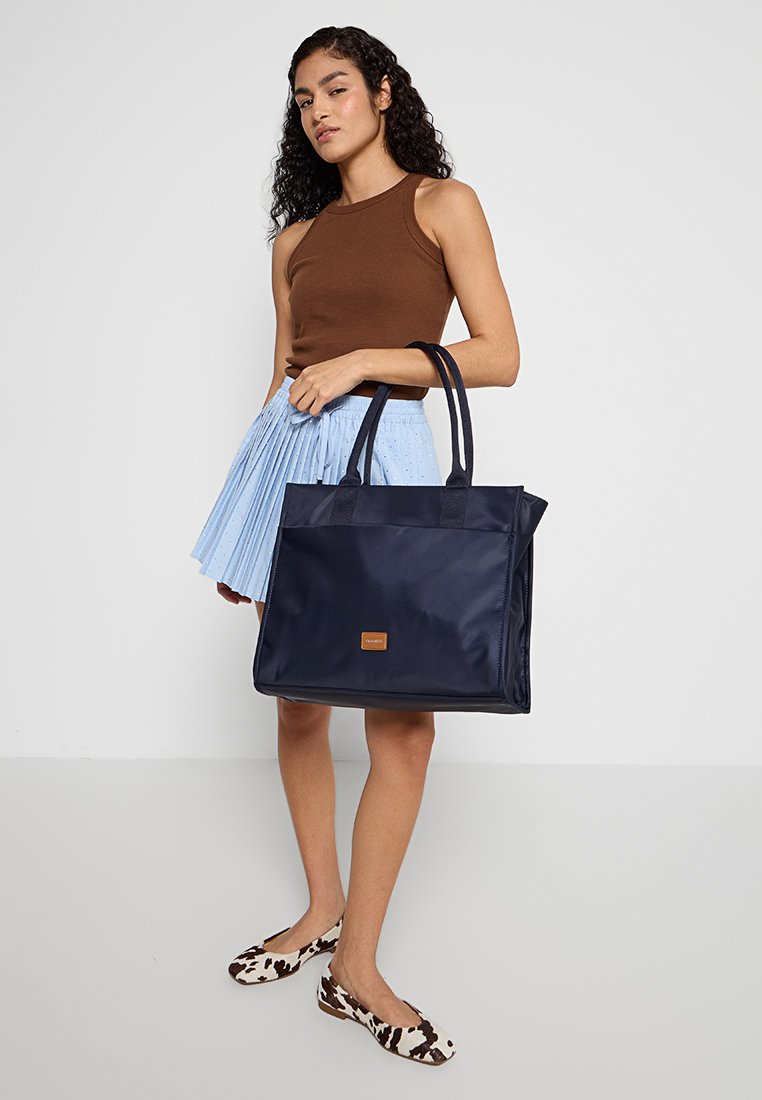 Marineblaues Nylon-Totebag mit zwei Griffen, rechteckiger Form und einem strukturierten braunen Etikett. Kombiniert mit einem plissierten hellblauen Rock und bedruckten Schuhen.