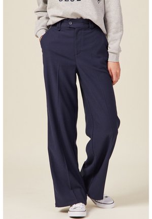 Personne portant un pantalon large bleu marine, un sweat-shirt gris et des baskets blanches, debout les mains dans les poches sur un fond clair.