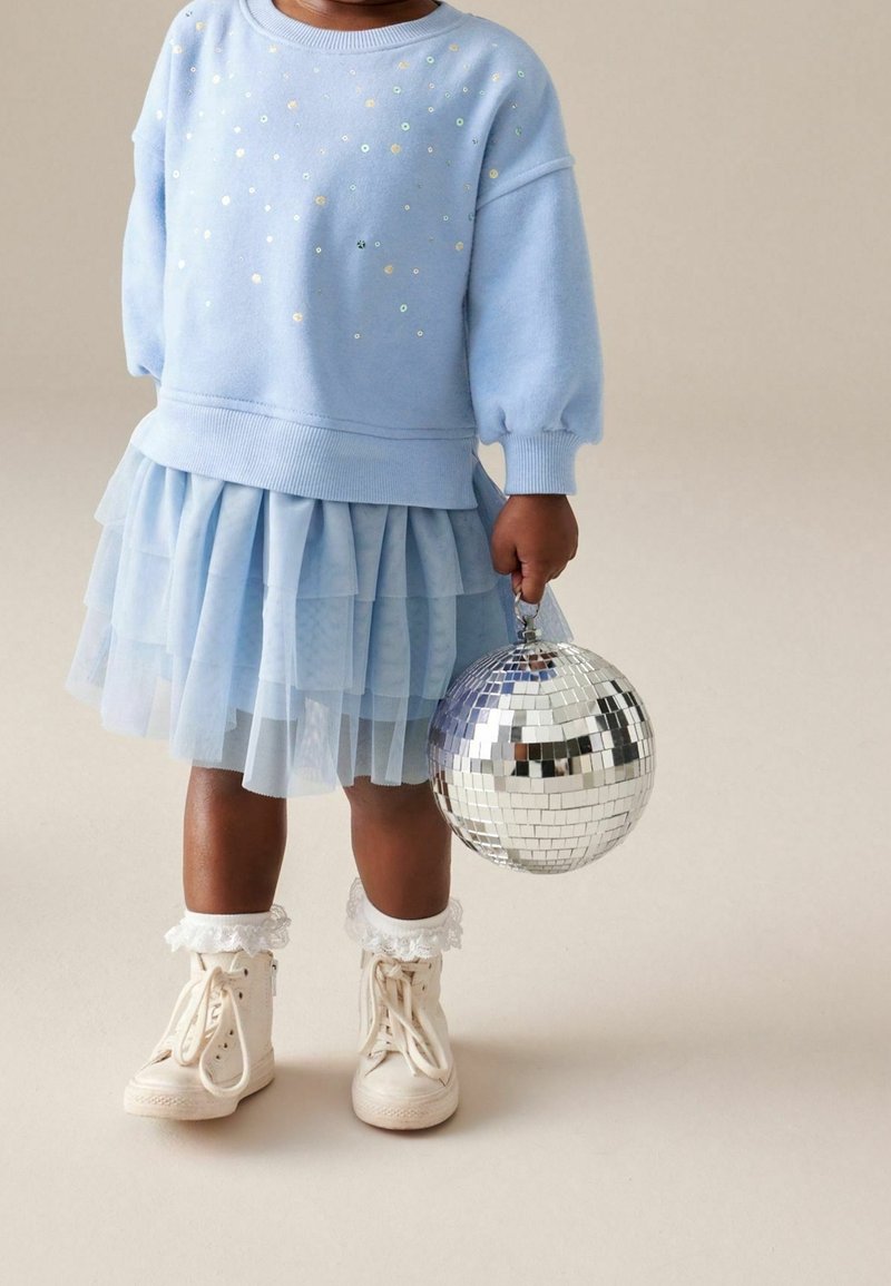 Enfant portant un pull bleu pailleté, une jupe en tulle bleue à volants, des baskets montantes blanches avec des chaussettes à lacets, tenant une boule disco argentée.