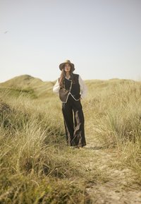 Gilet marron avec des bordures blanches sur une chemise à manches longues blanche, associé à un pantalon large noir et un chapeau beige, dans un paysage herbeux.
