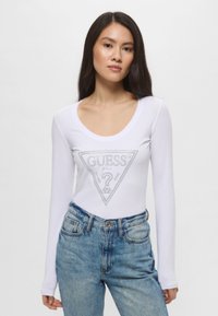 Maglietta bianca a maniche lunghe con un design del logo triangolare argentato al centro. Abbinata a jeans blu sbiaditi a vita alta.