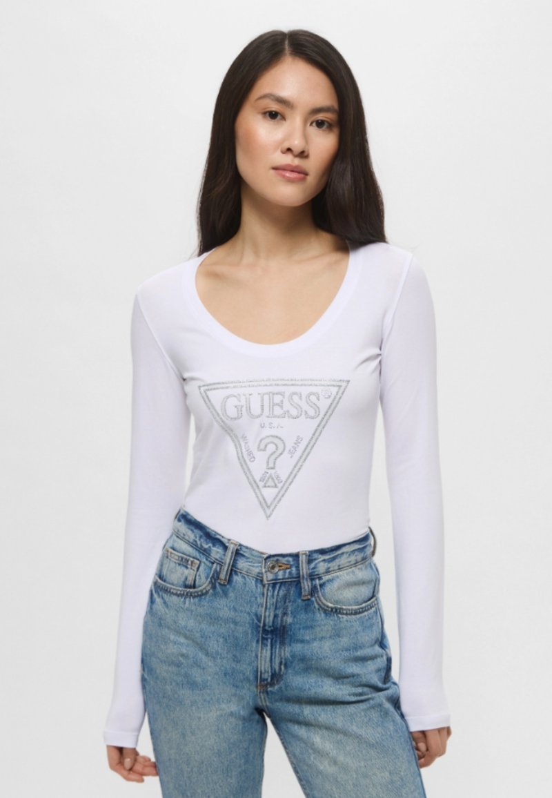 Maglietta bianca a maniche lunghe con un design del logo triangolare argentato al centro. Abbinata a jeans blu sbiaditi a vita alta.