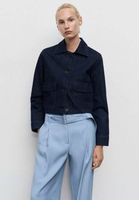 Dunkelblaue Jeansjacke mit Kragen, vorderem Knopfverschluss und zwei Brusttaschen, kombiniert mit hellblauen hochgeschnittenen Hosen.