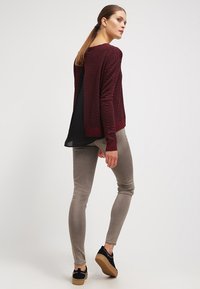 Maglione lavorato a maglia bordeaux con texture a zigzag, schiena nera a strati, abbinato a pantaloni grigi chiari slim-fit e scarpe nere con motivo.