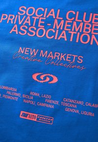 Blå stof t-shirt med pink grafisk tekst. Indeholder "SOCIAL CLUB PRIVATE-MEMBER ASSOCIATION" og forskellige italienske bynavne.
