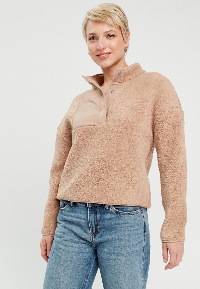 Pull en polaire beige avec un col zippé à mi-hauteur, surface texturée et une poche poitrine en toile beige ; associé à un jean bleu.