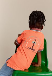 Koraalkleurig polo-shirt met een contrasterende kraag, voorzien van een grafische print van een palmboom en tekst, gecombineerd met blauwe jeans op een groene stoel.