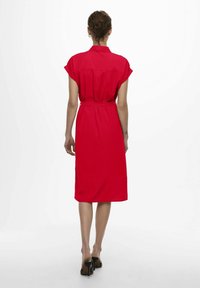 Robe rouge à manches courtes, col chemise, taille cintrée et longueur au genou. Style minimaliste sans motifs ni éléments métalliques visibles.